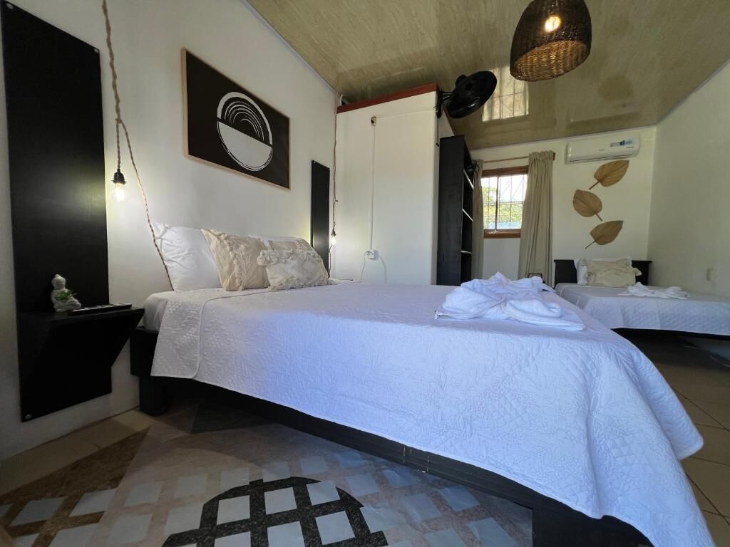 A bedroom with a bed, bedside tables, a hanging light fixture, a wall-mounted air conditioner, and a window with curtains.