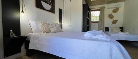 A bedroom with a bed, bedside tables, a hanging light fixture, a wall-mounted air conditioner, and a window with curtains.