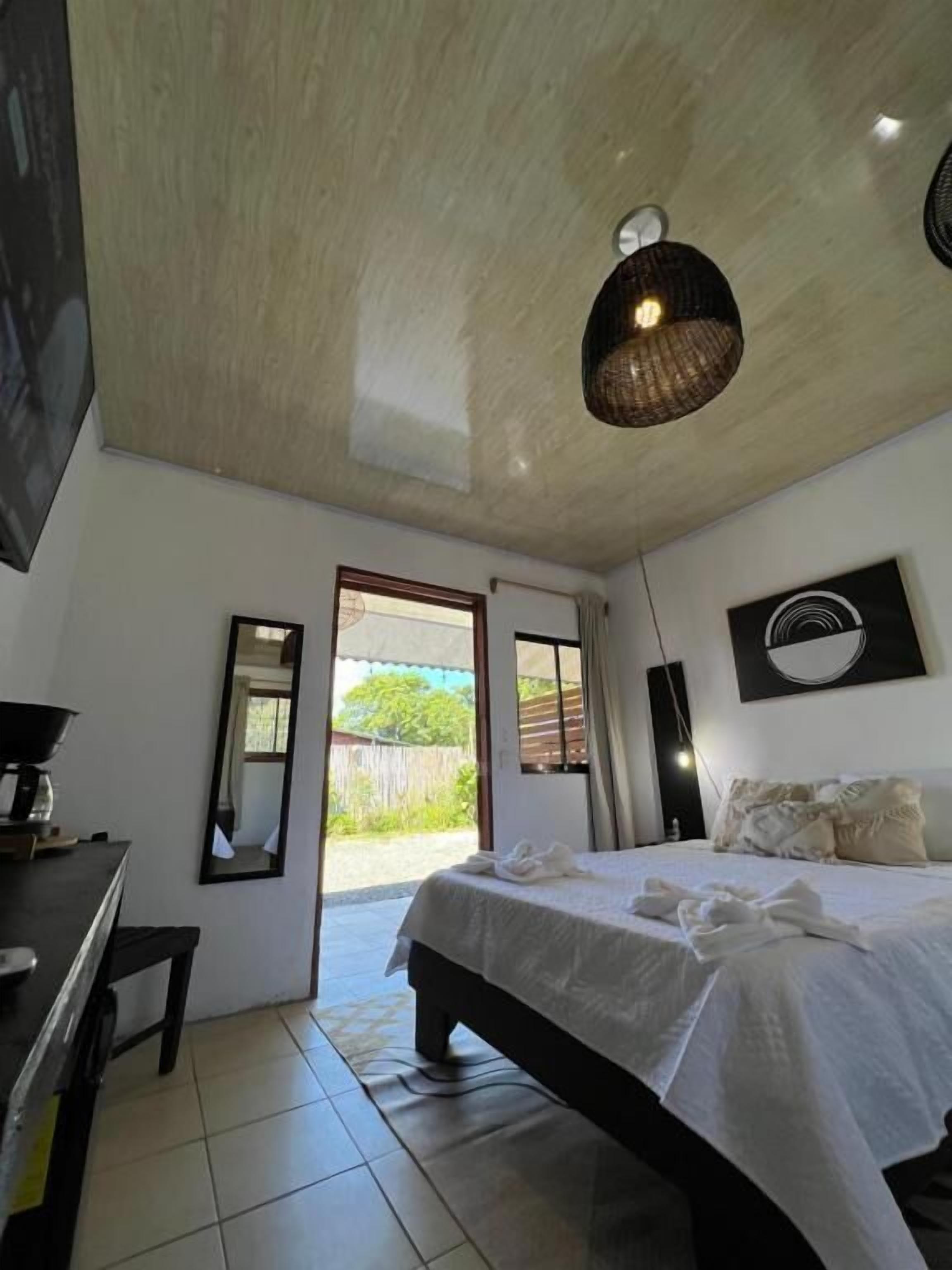 A bedroom with a bed, a desk, a mirror, a ceiling light fixture, and a view of the outdoors.