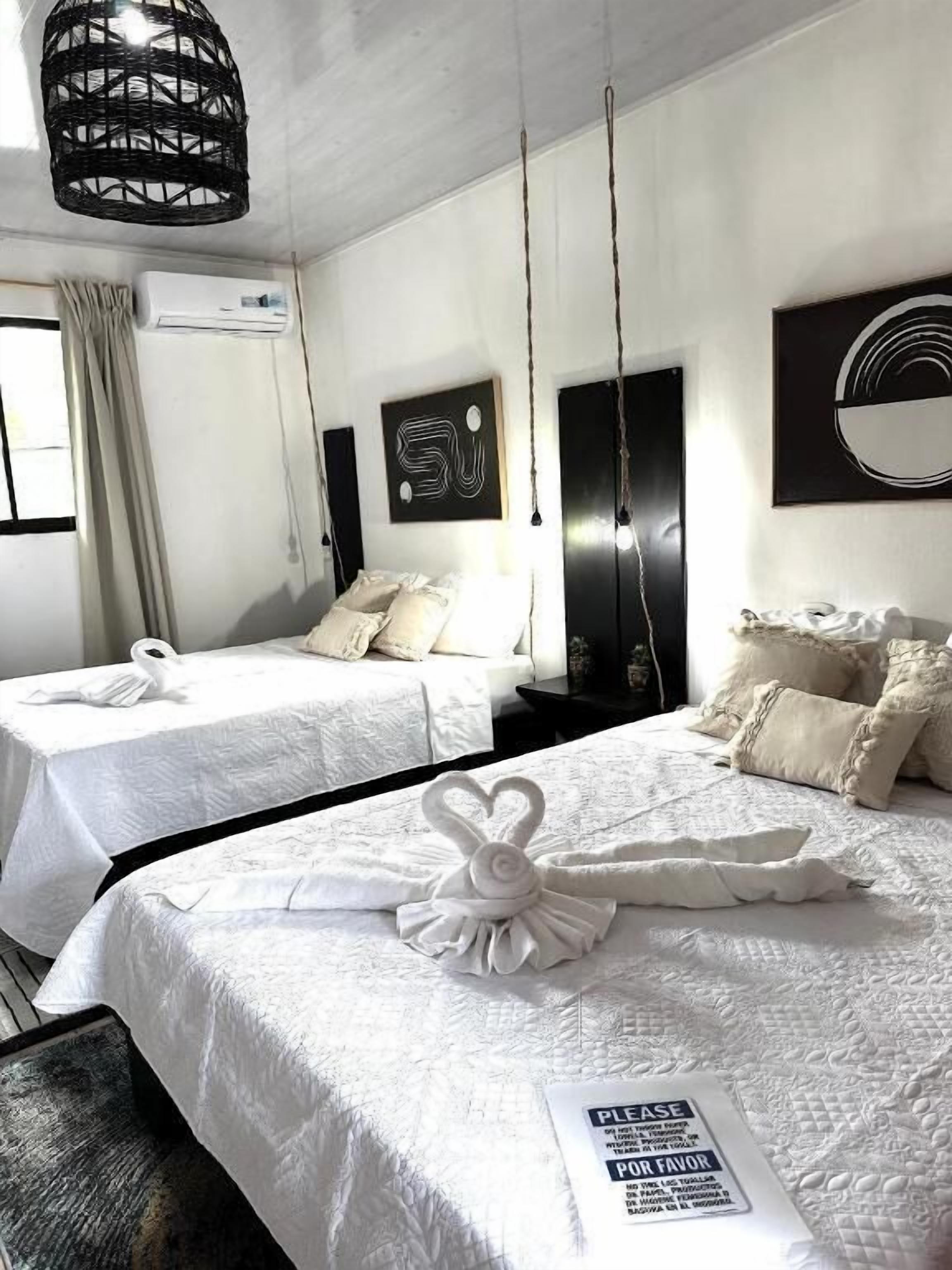 A hotel room with two beds, a hanging light fixture, a wall-mounted artwork, and a sign on the bed.