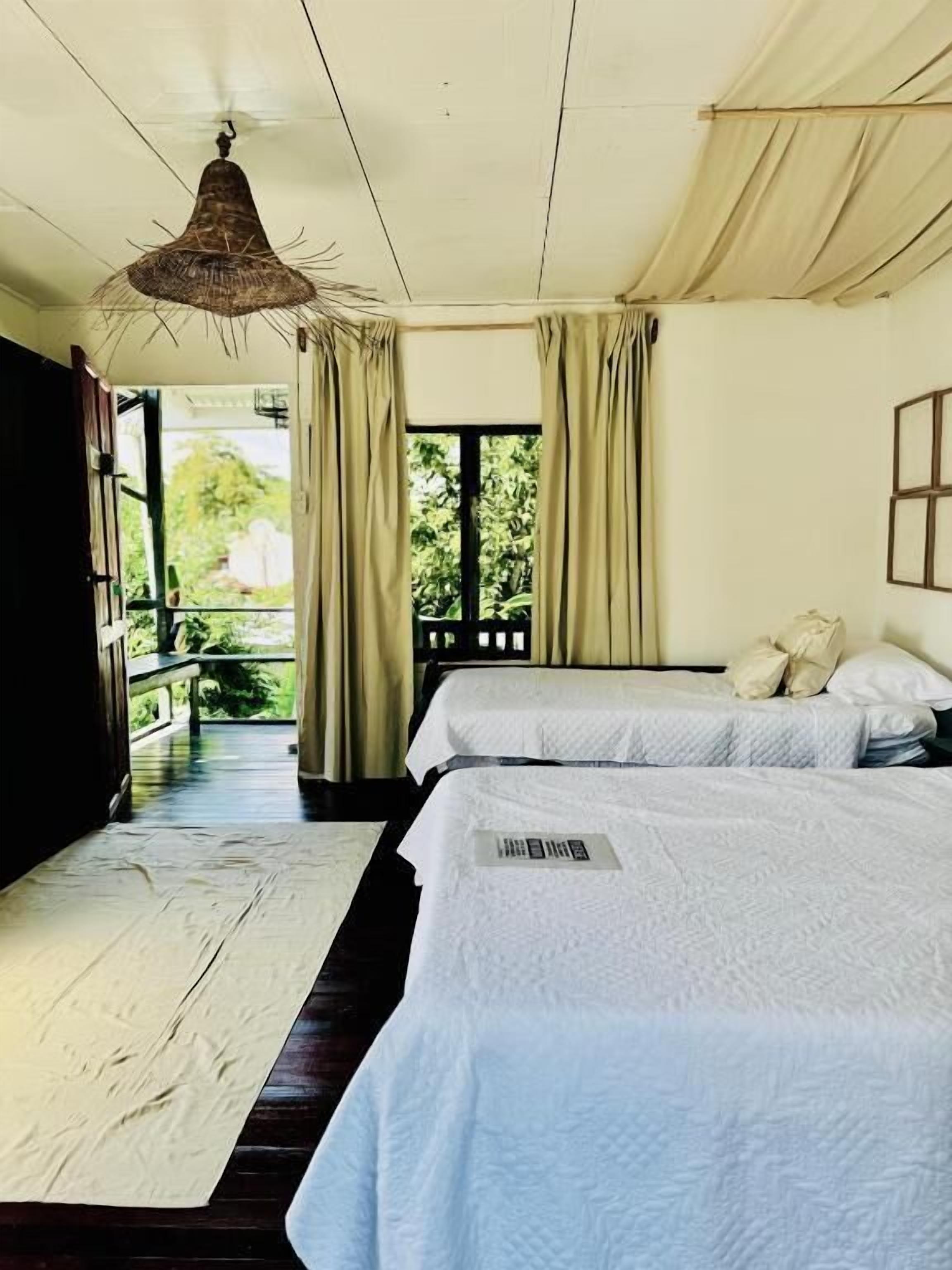 A room with two beds, a hanging light fixture, a window with curtains, and a view of greenery outside.