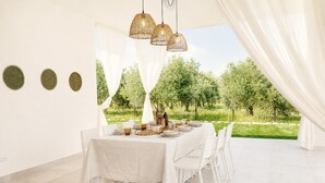 Un coin repas avec une table dressée pour quatre personnes, des chaises blanches et une vue sur la verdure à l’extérieur.