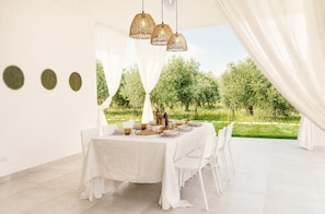 Un coin repas avec une table dressée pour quatre personnes, des chaises blanches et une vue sur la verdure à l’extérieur.