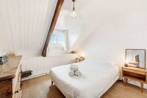 Ein Schlafzimmer mit schräg verlaufender Decke, einem Bett mit zusammengerollten Handtüchern, einem Nachttisch mit Lampe und einem Fenster.