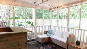 A covered outdoor seating area with a wooden bench, a cushioned bench with a throw blanket, and a wicker table.