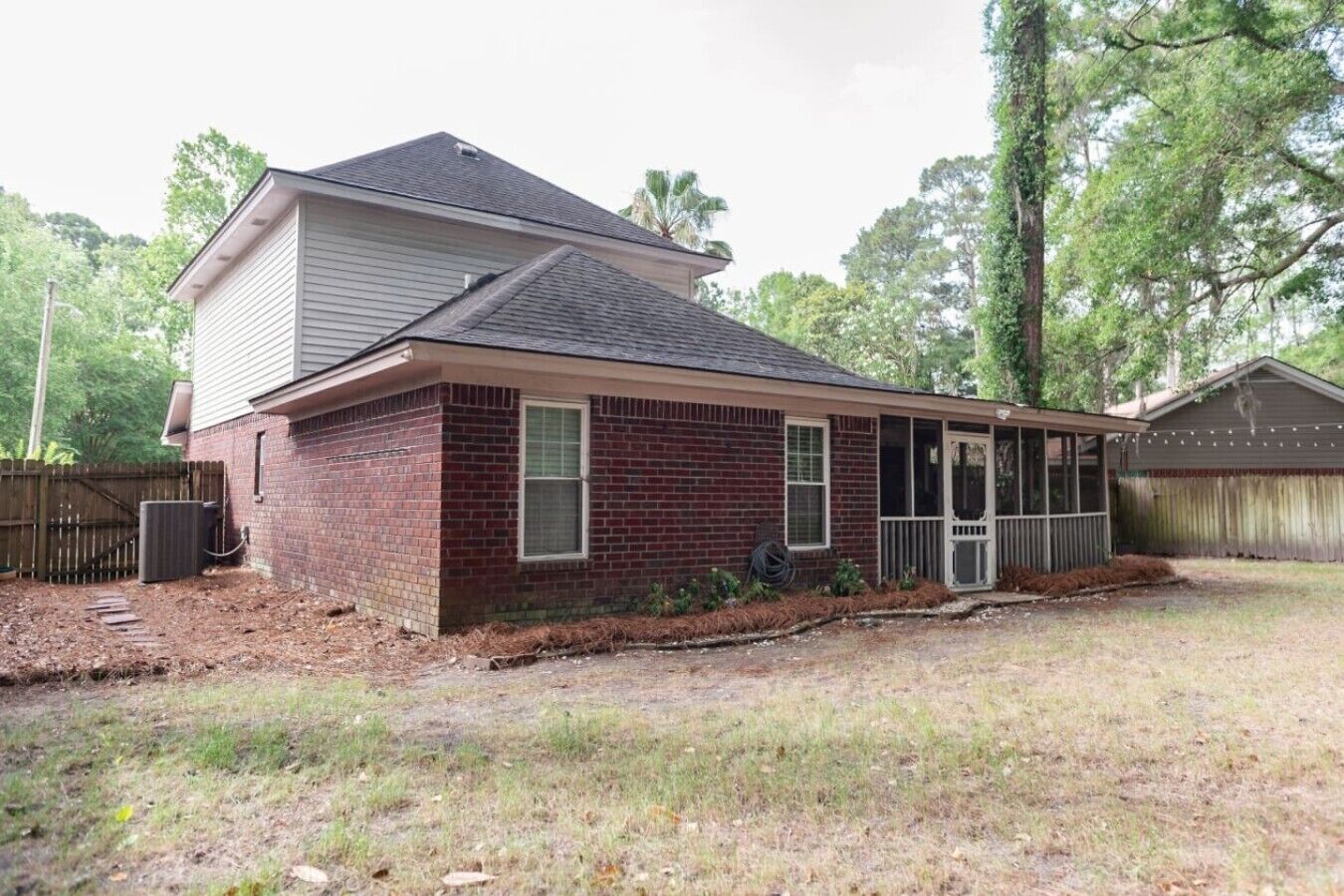 A single-story brick house with a covered porch, a fenced yard, and trees in the background.