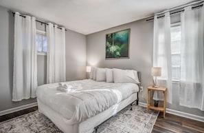 A bedroom with a bed, a nightstand, a lamp, a painting, and a window with curtains.