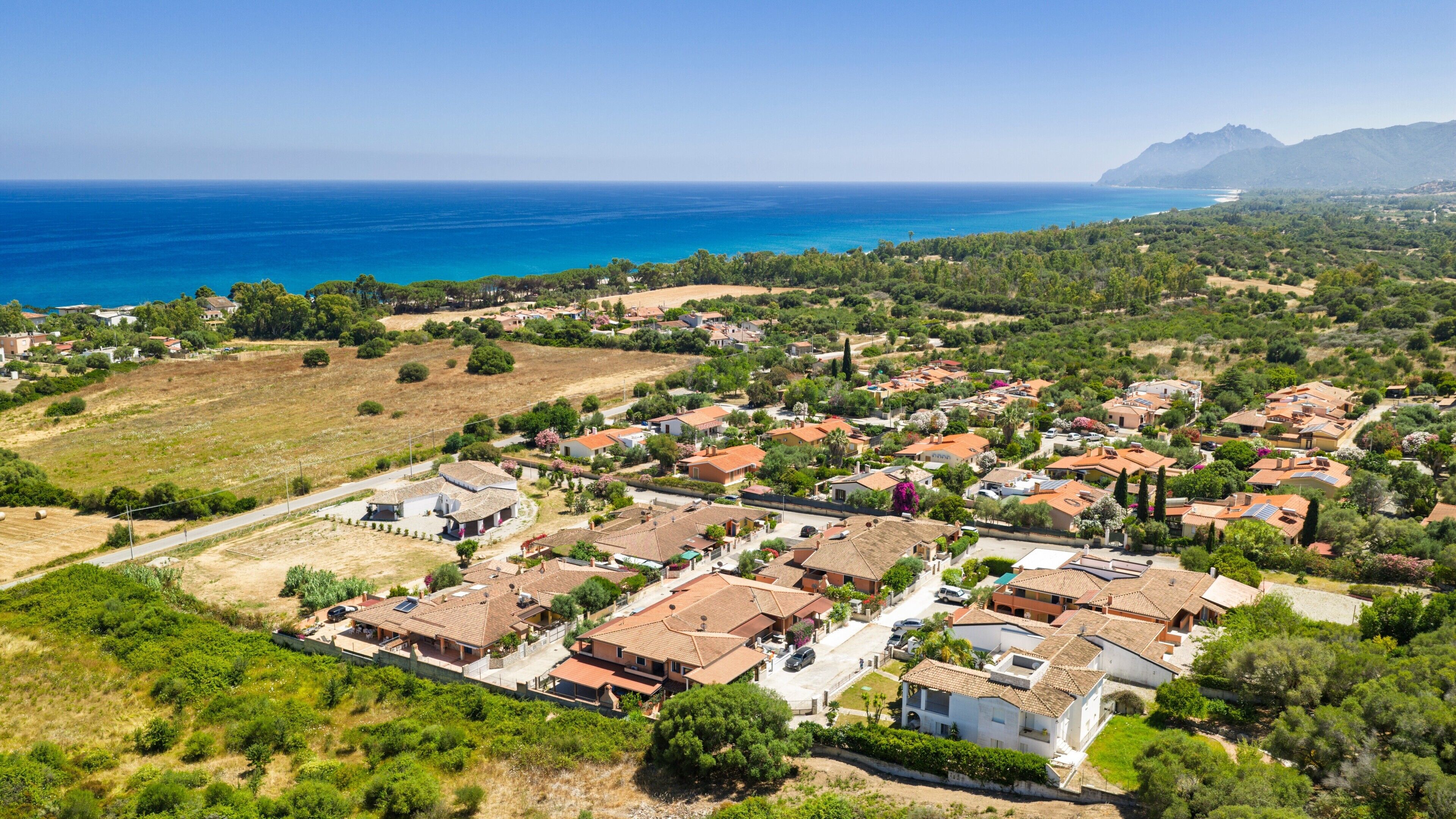 Vue aérienne d’un quartier résidentiel situé sur la côte, avec des maisons, des routes et une vue dégagée sur la mer et les montagnes au loin.