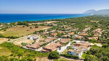 Vue aérienne d’un quartier résidentiel situé sur la côte, avec des maisons, des routes et une vue dégagée sur la mer et les montagnes au loin.