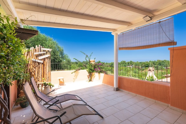 Eine überdachte Terrasse mit Liegestühlen, Topfpflanzen und Blick auf Grünflächen sowie ein entferntes Gebäude.