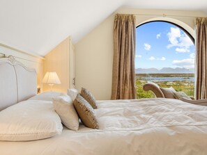 Ein Schlafzimmer mit einem großen Bett, einem Fenster mit Vorhängen, einer Lampe und Blick auf eine Landschaft.