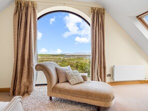Ein Zimmer mit einem gebogenen Sessel, beigen Vorhängen und Blick auf eine Landschaft durch ein großes Bogenfenster.