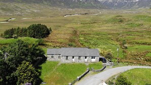 Une maison en pierre avec un toit en pente, entourée d’un paysage rural avec des montagnes en arrière-plan.