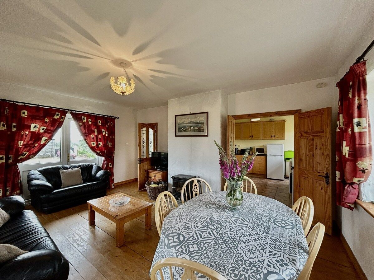 Un salon chaleureux avec un canapé en cuir noir, une table basse en bois et une table à manger avec des chaises. On peut apercevoir la cuisine à travers une porte ouverte.