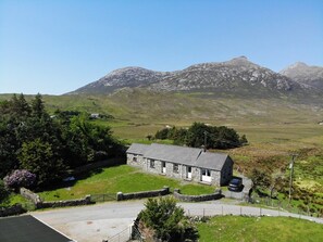 A stone house with a car parked in front, surrounded by a stone wall and greenery, with mountains in the background. - Twelve Bens Lodge (Recess)