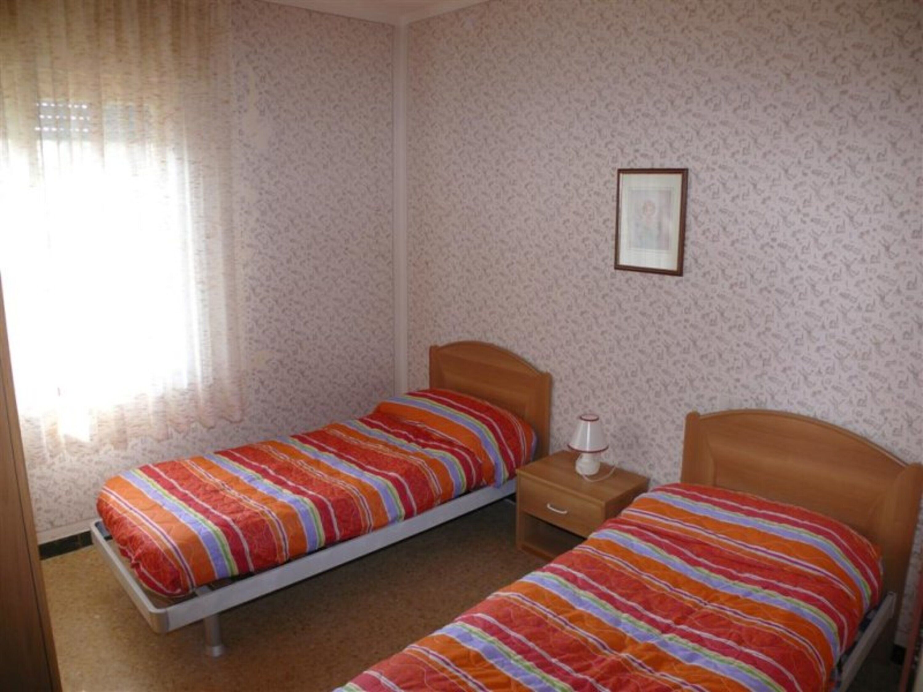 A small bedroom with two single beds, a nightstand, a framed picture on the wall, and a window with curtains.