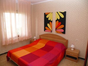 Ein Schlafzimmer mit einem Bett, zwei Nachttischen, einem Fenster mit Vorhängen und zwei an der Wand befestigten Kunstwerken.