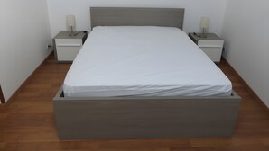 Un lit simple avec une tête de lit et un pied de lit en bois, flanqué de deux tables de chevet avec des lampes, dans une pièce au plancher en bois et aux murs blancs.