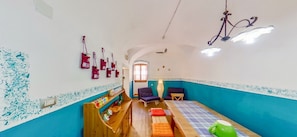 Una habitación con cama, dos sillas, una estantería con botellas, una lámpara colgante y una ventana.
