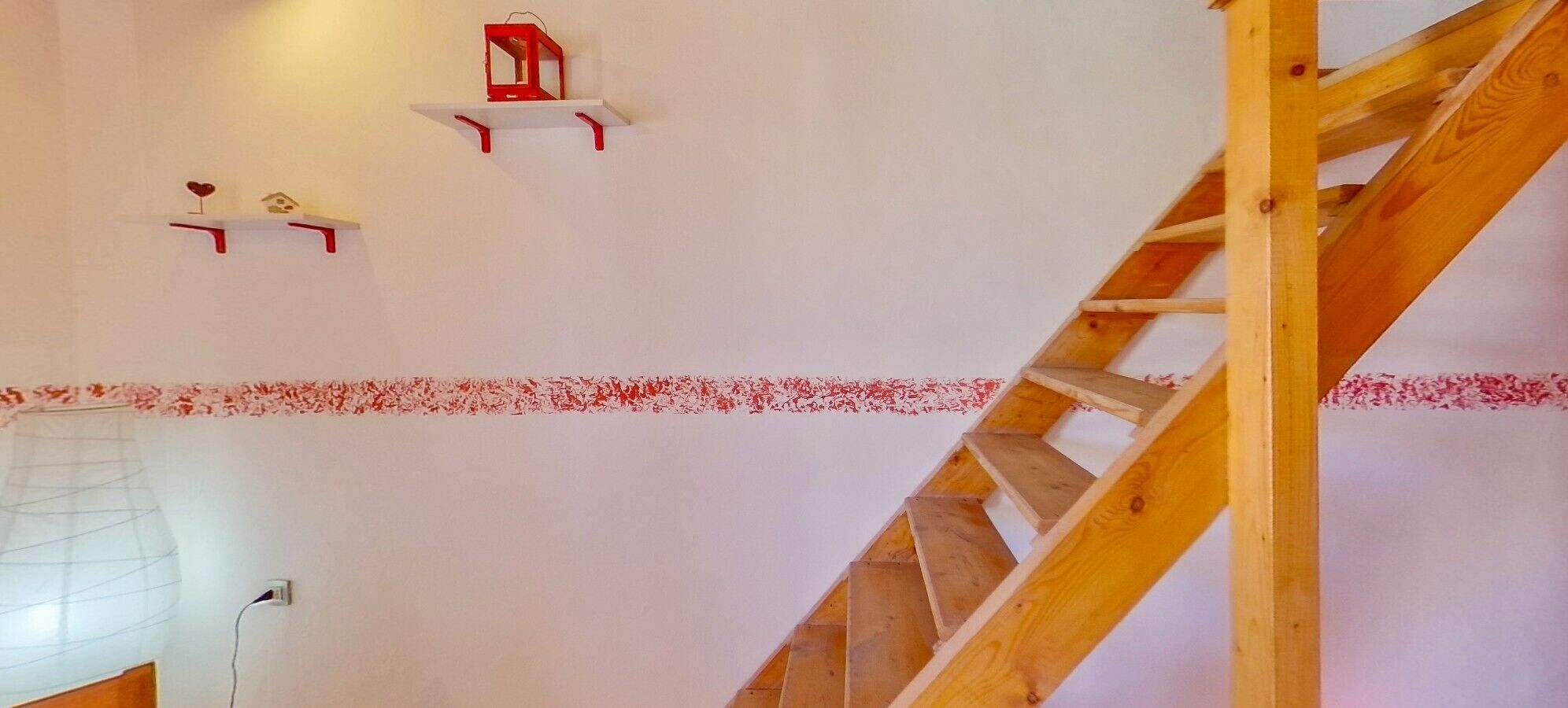 Un escalier avec des marches en bois et une rampe, un mur blanc orné d’une bordure florale rouge, et deux étagères rouges décorées de motifs en forme de cœur.