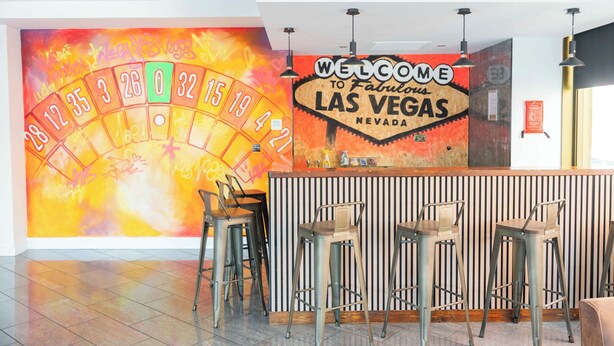 Eine Casino-Lobby mit Wandmalerei eines Roulette-Spiels, einem Schild mit der Aufschrift 'Welcome to Fabulous Las Vegas Nevada' und Barhocker.