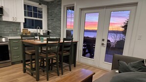 A modern kitchen with a dining table and chairs, a bench, and a view of a sunset through a glass door. - Stunning Bluff Cottage with Panoramic Puget Sound & Olympic Mountain Views (Seattle)
