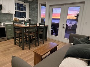 A modern kitchen with a dining table and chairs, a bench, and a view of a sunset through a glass door. - Stunning Bluff Cottage with Panoramic Puget Sound & Olympic Mountain Views (Seattle)