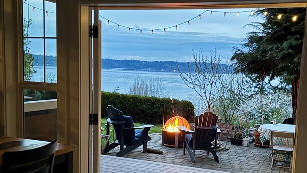 A view from a room to an outdoor deck with a fire pit, chairs, and a table. - Stunning Bluff Cottage with Panoramic Puget Sound & Olympic Mountain Views (Seattle)