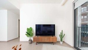 Moderni olohuone, jossa on litteänäyttöinen televisio puisen konsolipöydän päällä, kaksi ruukkukasvia ja näkymä parvekkeelle lasiovien läpi.