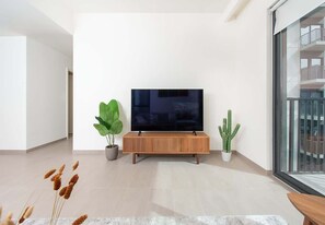 Ein modernes Wohnzimmer mit einem Flachbildfernseher auf einer Holzkonsole, zwei Topfpflanzen und Blick auf einen Balkon durch eine Glastür.