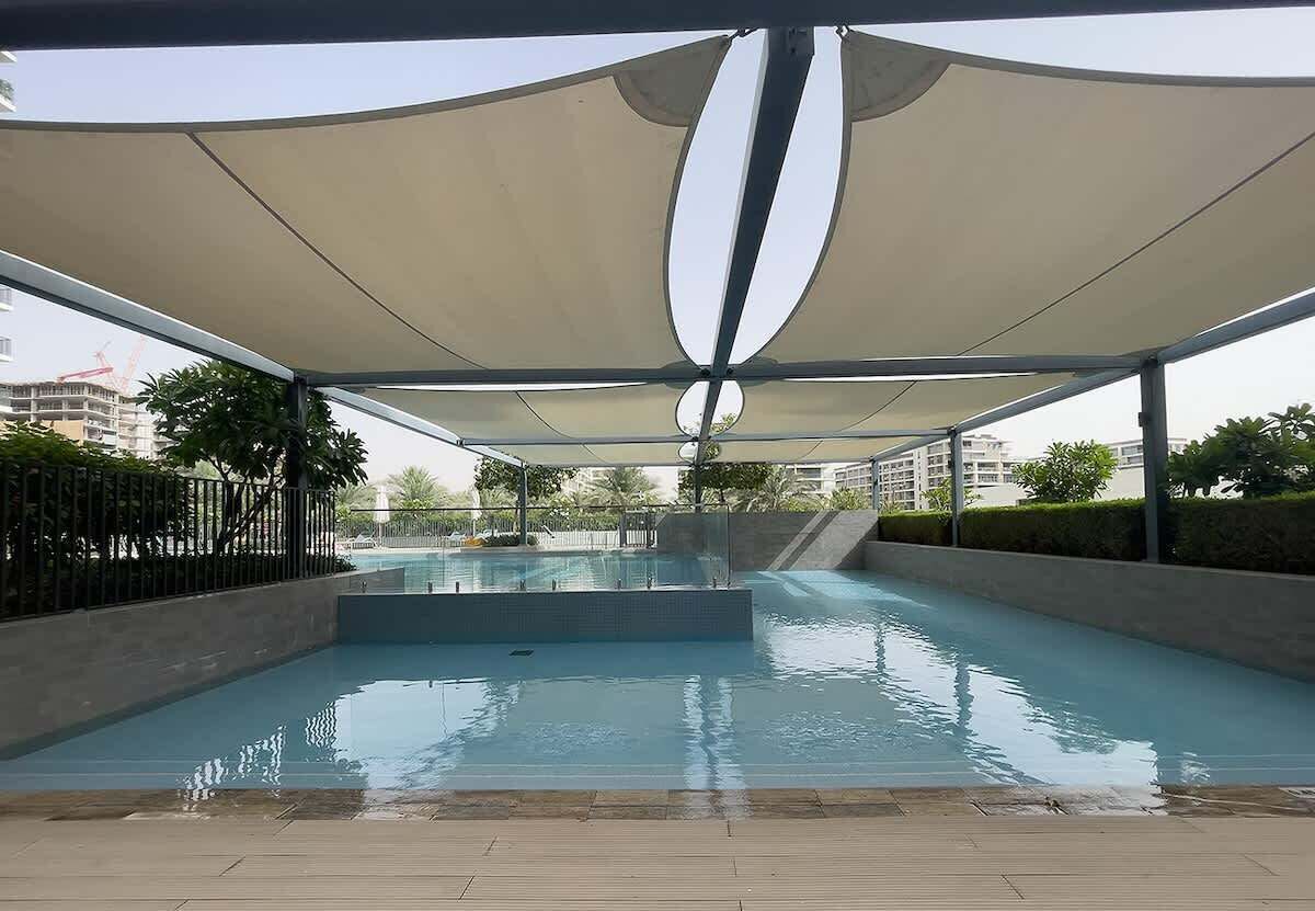 A swimming pool with a shaded area overhead, surrounded by a low fence and greenery.