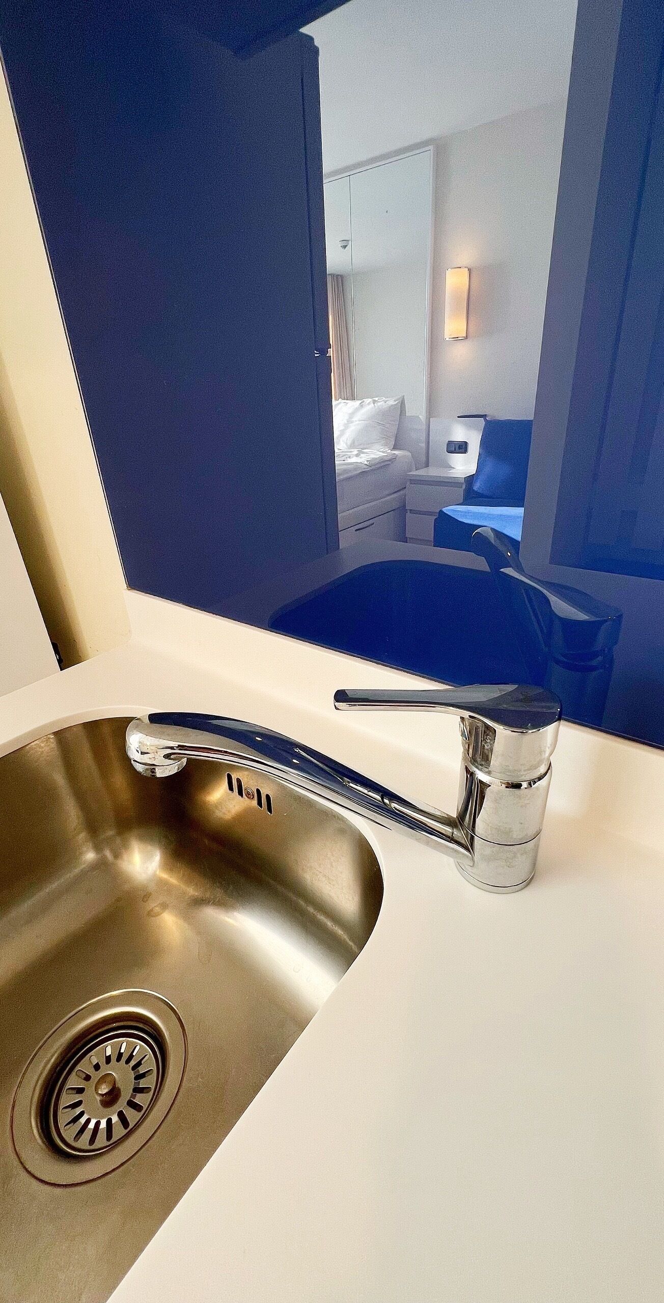Un baño de hotel moderno con un lavamanos de acero inoxidable, un espejo y vistas al dormitorio contiguo.