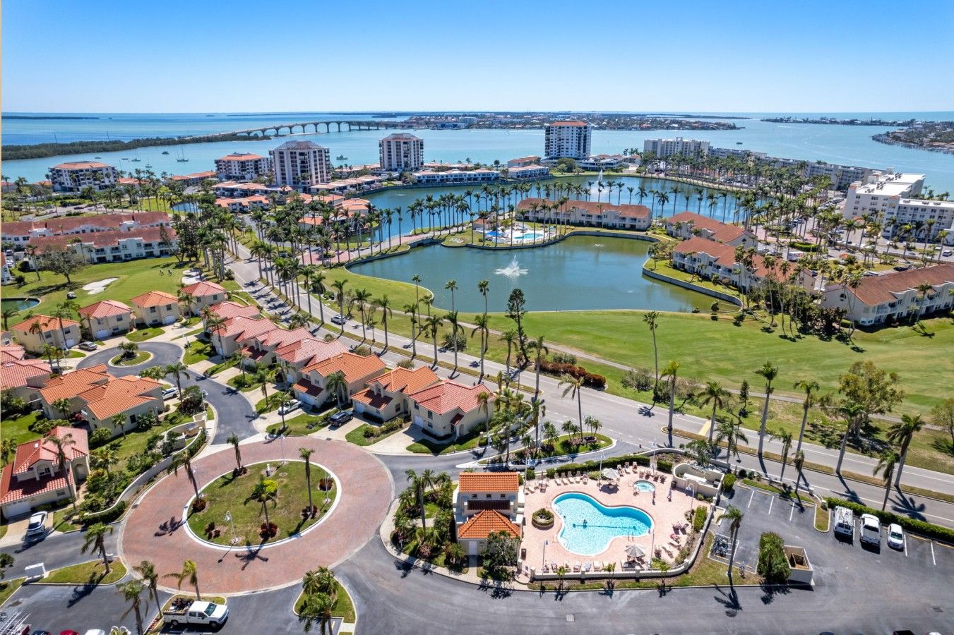 A resort with a central swimming pool, connected to a marina, surrounded by residential buildings and palm trees.