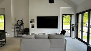Een moderne woonkamer met een grote grijze bank, een flatscreen televisie aan de muur en een groot raam met uitzicht op groen.