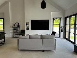 Ein modernes Wohnzimmer mit einem großen grauen Sofa, einem flachbildfernseher an der Wand und einem großen Fenster mit Blick auf Grünfläche.