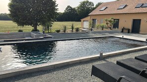 Ein Swimmingpool mit umgebender Liegefläche, ein Gebäude mit Ziegeldach und ein großer Baum.