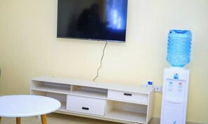 Une piĂšce avec un tĂ©lĂ©viseur fixĂ© au mur, un meuble de rangement blanc, une petite table ronde et un distributeur dâeau.