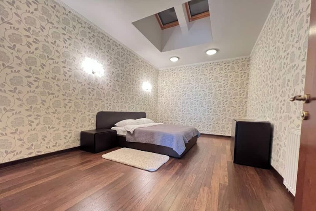 Un dormitorio con cama, mesita de noche, alfombra, techo con tragaluces y papel tapiz estampado.