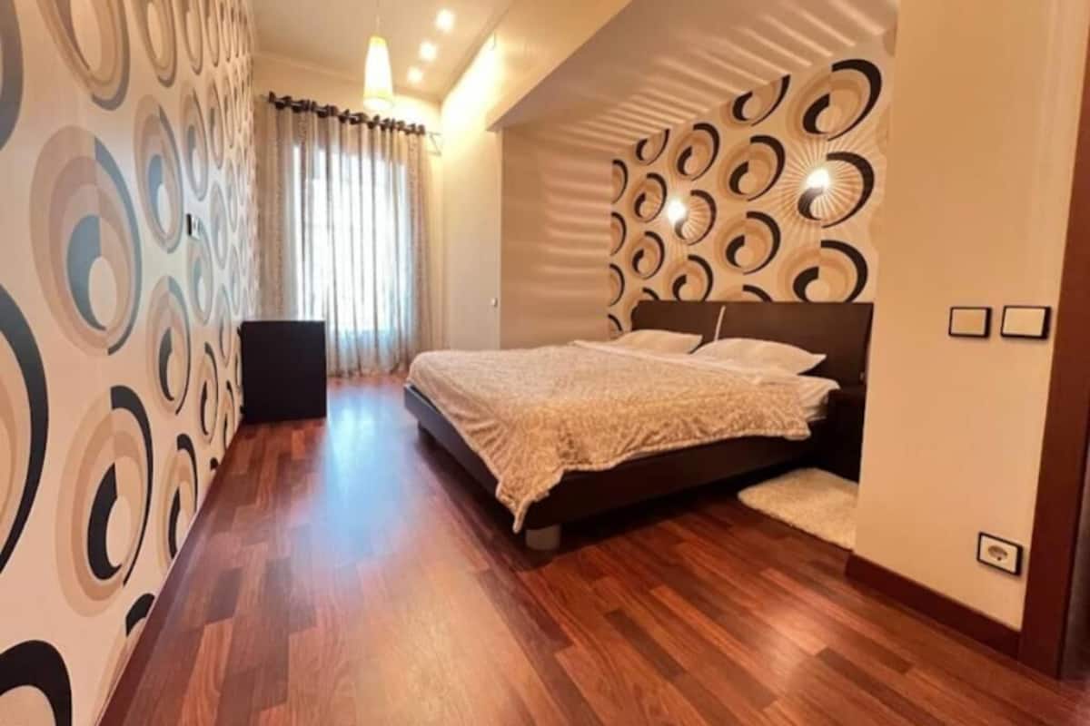 Un dormitorio con una cama, una pared con diseños circulares, una ventana con cortinas y suelo de madera.