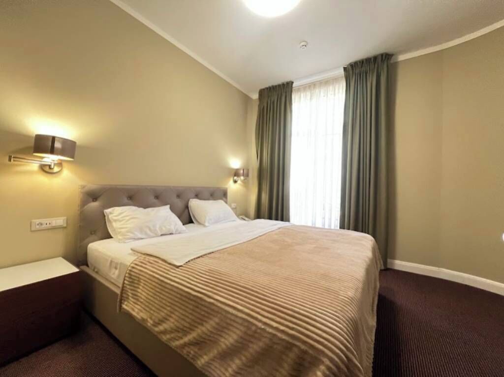 A hotel room with a bed, two bedside lamps, a nightstand, and a window with curtains.