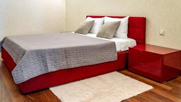 Una cama con cabecera roja, colcha gris y colchón blanco. Hay una mesita de noche roja con cajones y una alfombra blanca y mullida sobre el suelo de madera.