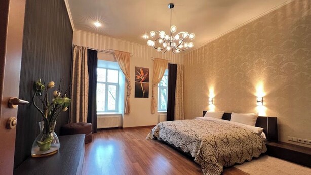 Un dormitorio con una cama grande, una araña y un florero con flores sobre una mesita de noche.
