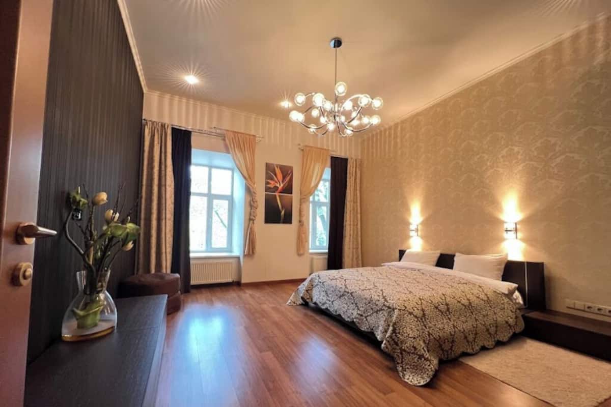 Un dormitorio con una cama grande, una araña y un florero con flores sobre una mesita de noche.