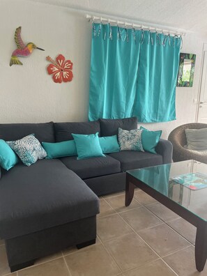 Ein Wohnzimmer mit einem Ecksofa, türkisfarbenen Kissen, einem Glastisch und einem Fenster mit türkisfarbenen Vorhängen.