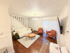 Ein Wohnzimmer mit einer Couch, zwei Sesseln, einem Couchtisch, einer Treppe und einem an der Wand befestigten Fernseher.