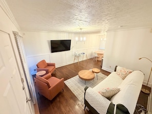 Ein modernes Wohnzimmer mit Fernseher, einer Couch, zwei Sesseln, einem Couchtisch und einem Essbereich mit Tisch und Stühlen.