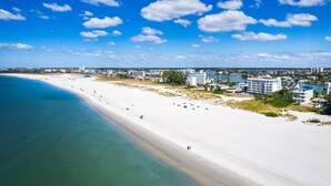 A beach with a white sandy shore, clear blue water, and a cityscape in the background. - Premium Corner Suite w/ Gulf Views | Beach & Pool (Treasure Island)