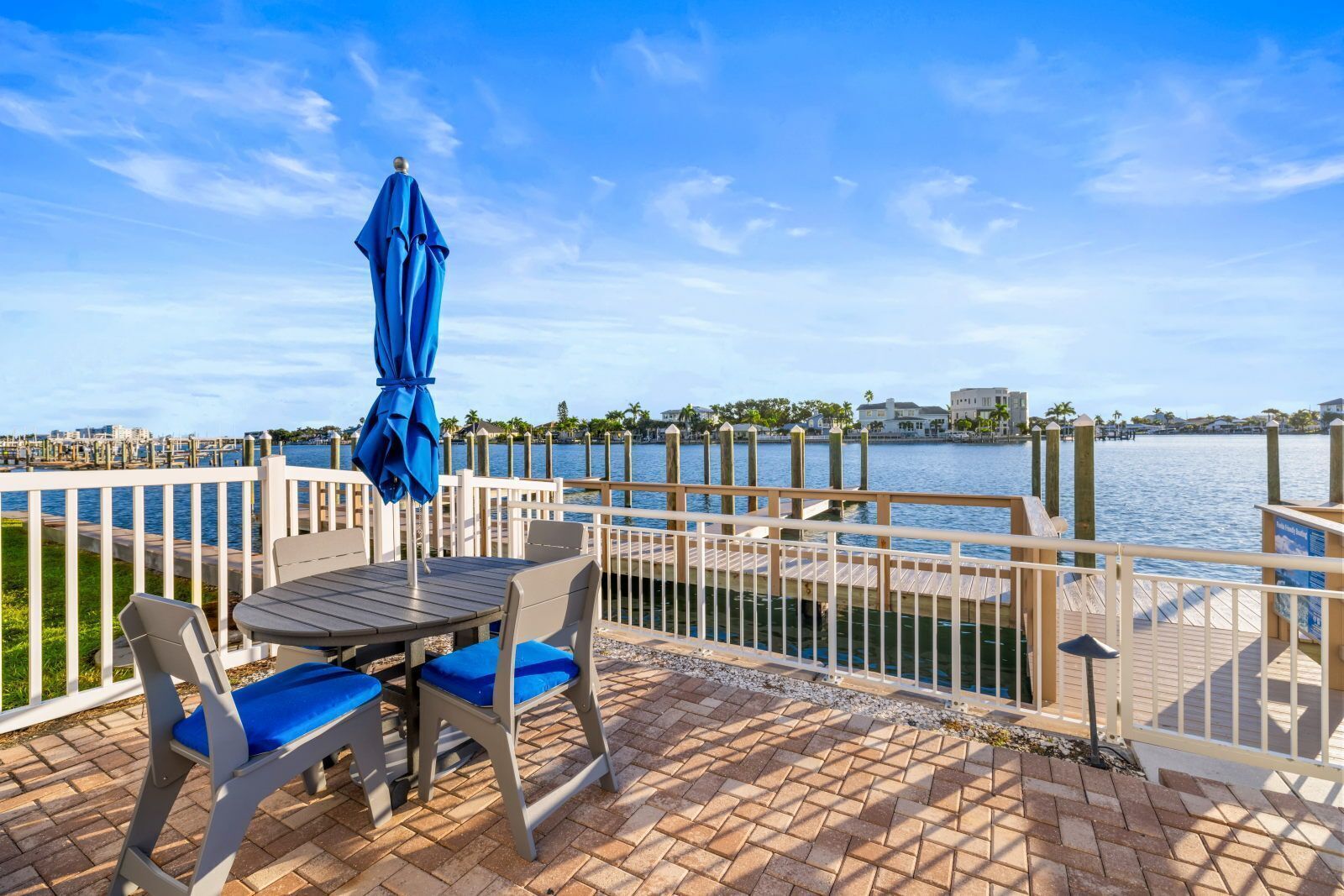 A waterfront patio with a table and chairs, a blue umbrella, and a view of the water and distant buildings.