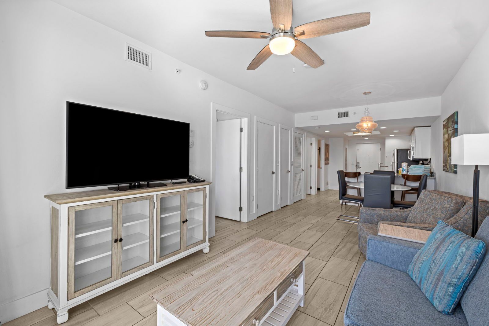 A modern living room with a flat-screen TV, wooden cabinet, ceiling fan, and a dining area with chairs and a table.