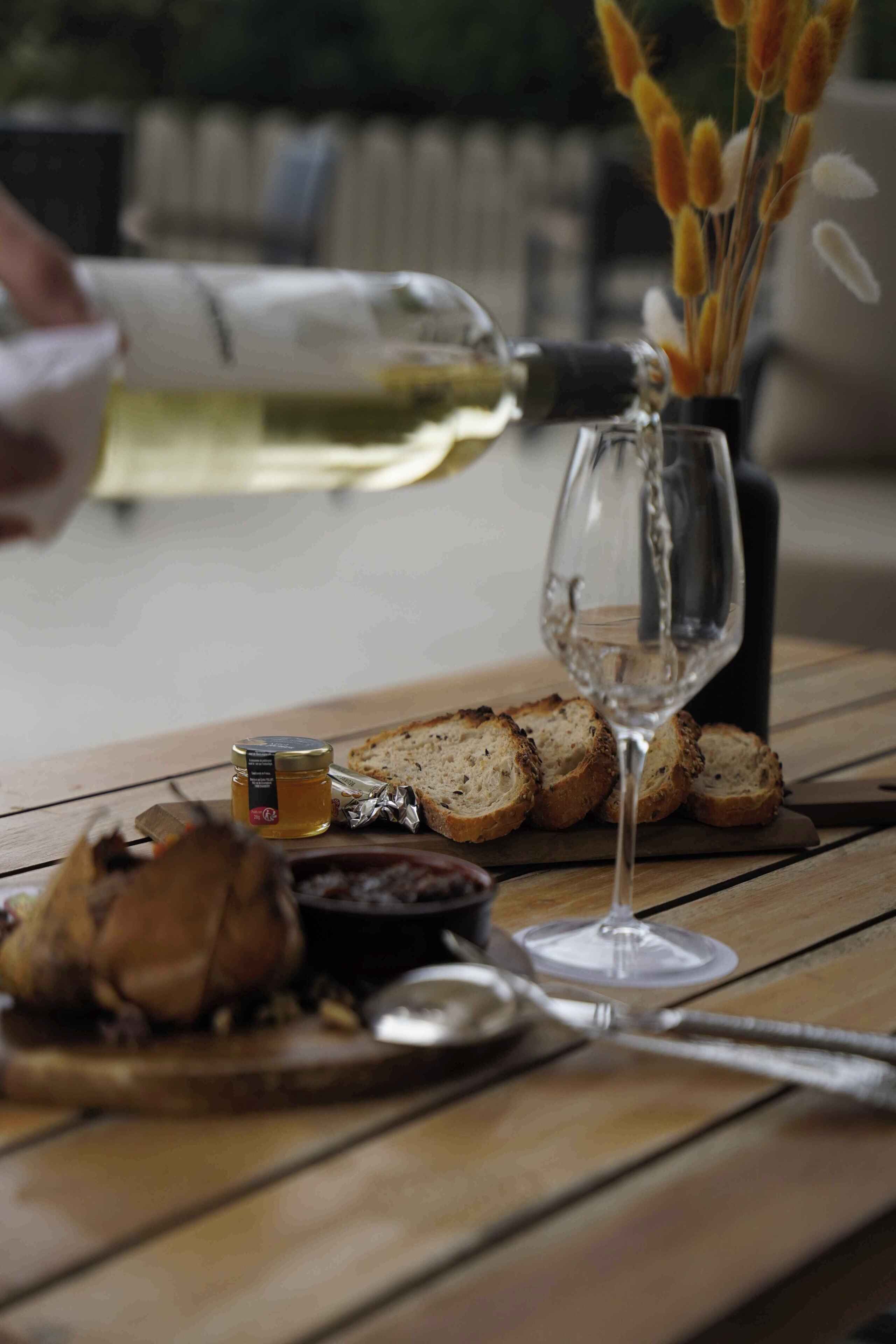 Une table en bois avec un verre de vin blanc, des tranches de pain, un pot de miel et un couteau.